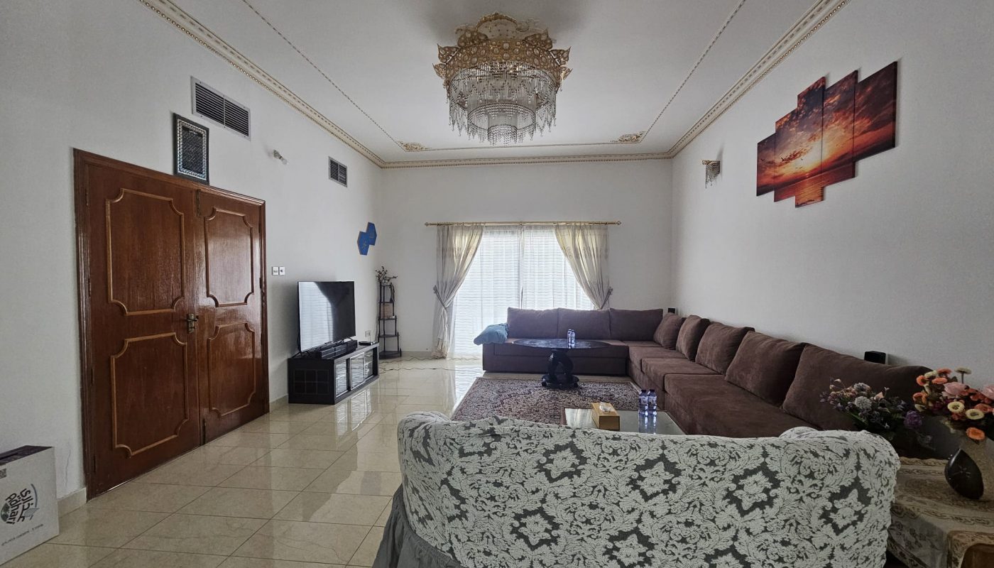 Spacious living room with a large L-shaped sofa, decorative ceiling light, TV, brown curtains, and a patterned slipcover on an armchair. Walls are adorned with artwork.
