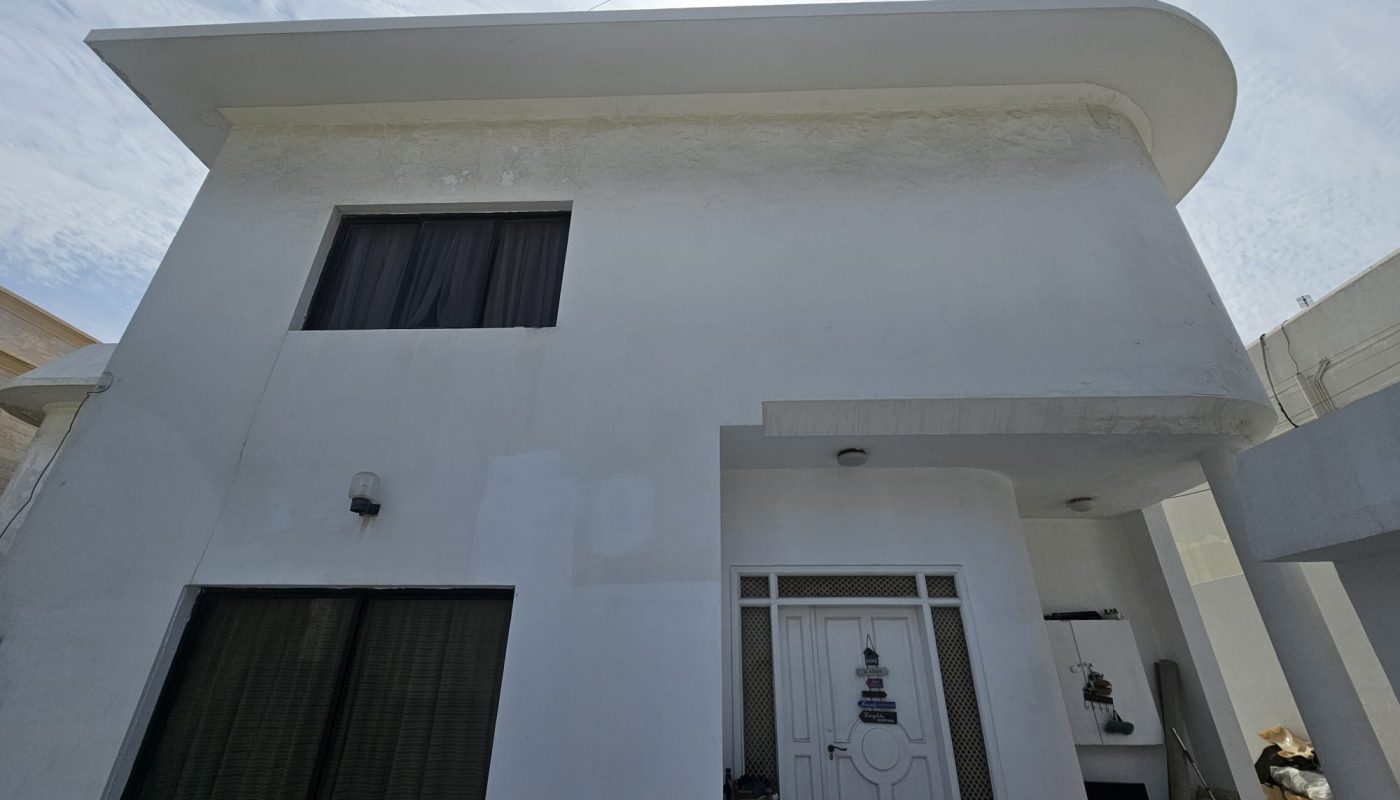 Modern two-story white building with large windows and a flat roof, featuring a closed white front door. A water dispenser is visible near the entrance.
