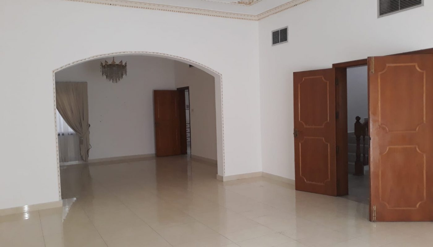 Spacious empty room with glossy tiled floors, white walls, and two wooden doors. A chandelier hangs from the ceiling, and an archway leads to another room with curtains.