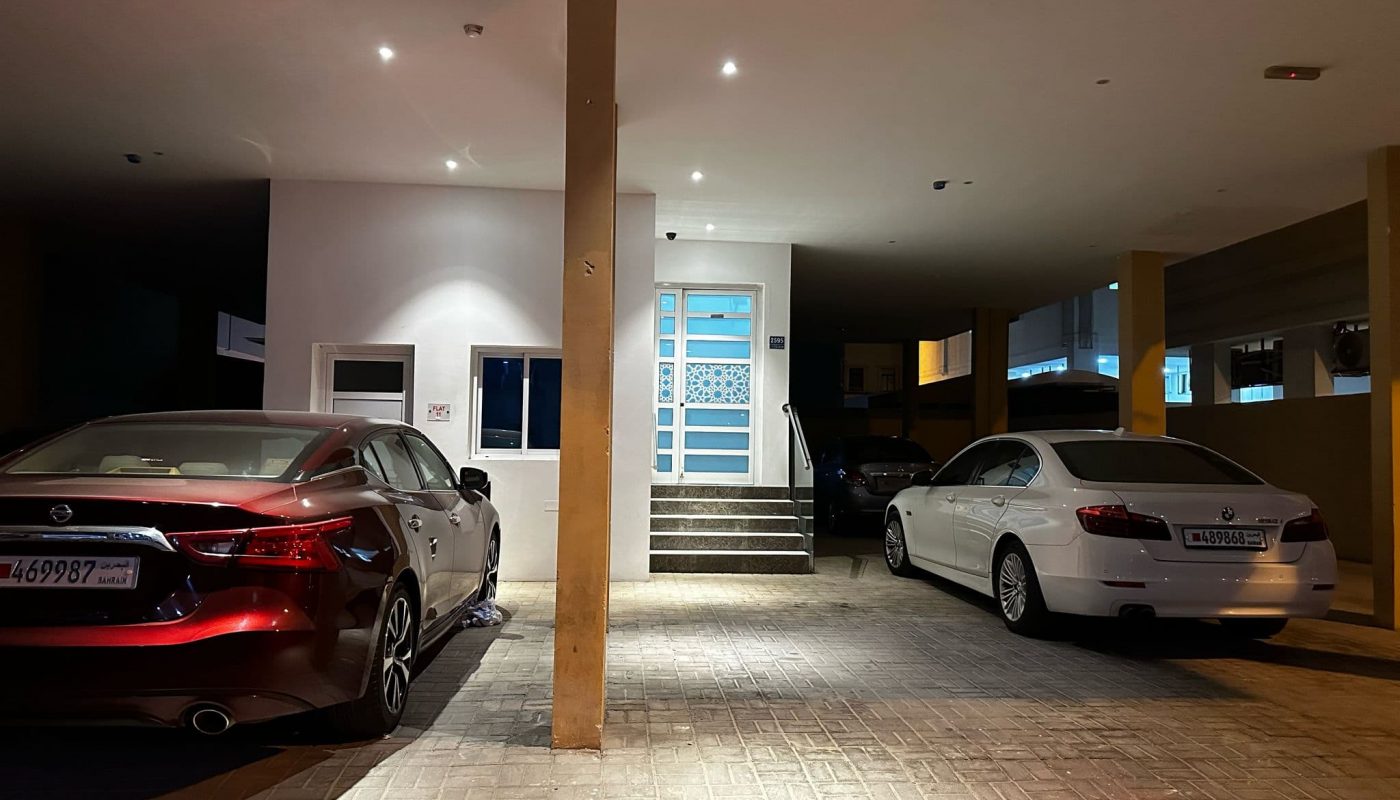 Covered parking area at night with a red car on the left, a white car on the right, and steps leading to a door in the background.