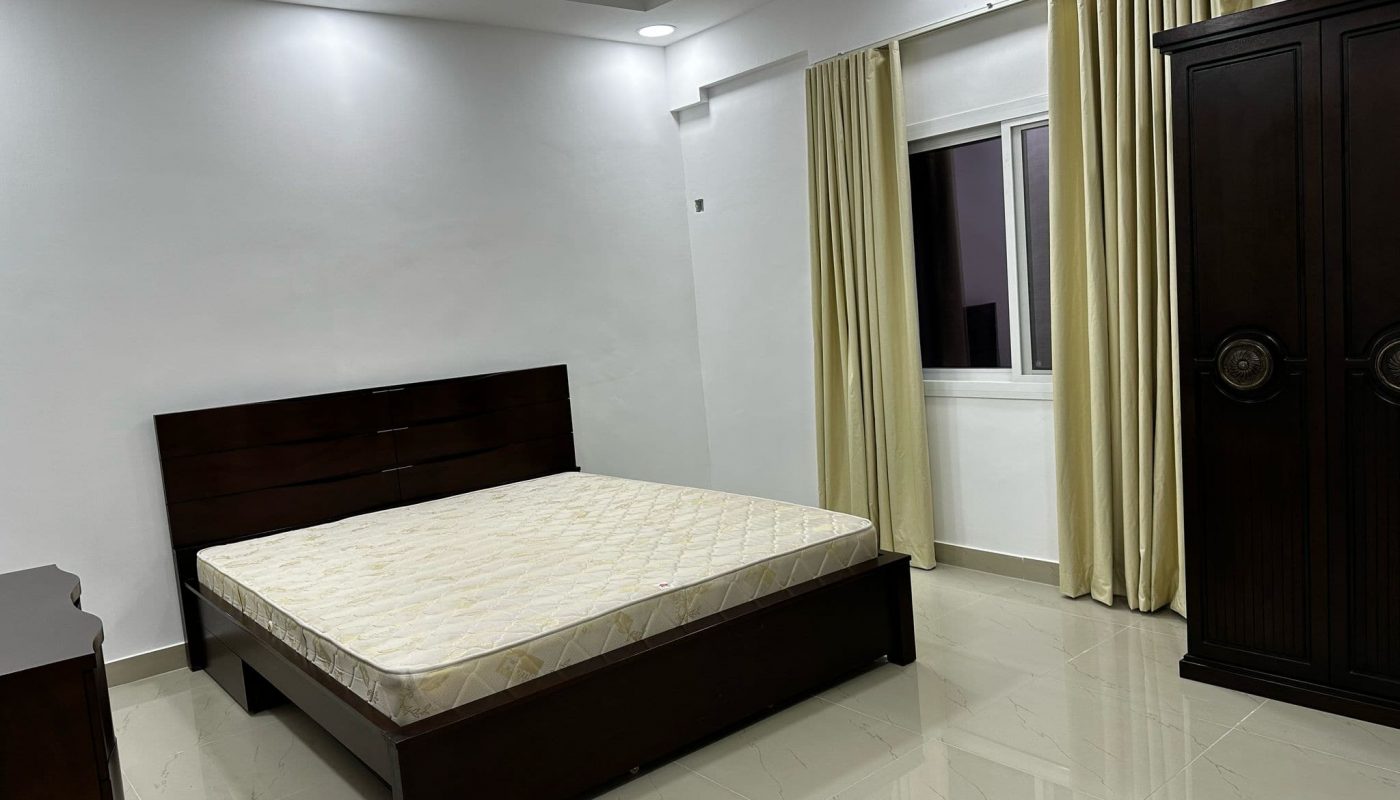 A minimalist bedroom with a dark wood bed frame and mattress, a wardrobe, cream curtains, tiled floor, and ceiling fan. The room is well-lit with recessed ceiling lights.