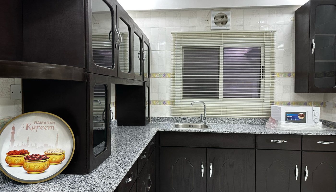 Modern kitchen with dark cabinets, granite countertops, a microwave, a decorative Ramadan Kareem plate, a window with blinds, and white tiled walls.