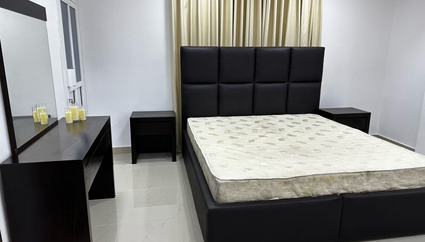 A modern bedroom with a large black upholstered bed frame, two black nightstands, a black dressing table with a mirror, candles, and beige curtains against a white wall.