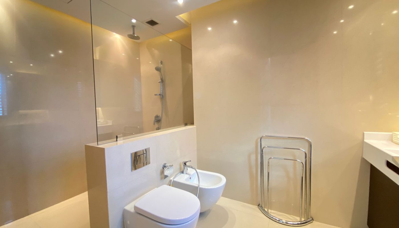 Modern bathroom with beige walls, featuring a wall-mounted toilet, bidet, walk-in shower with glass partition, towel rack, and countertop with sink.