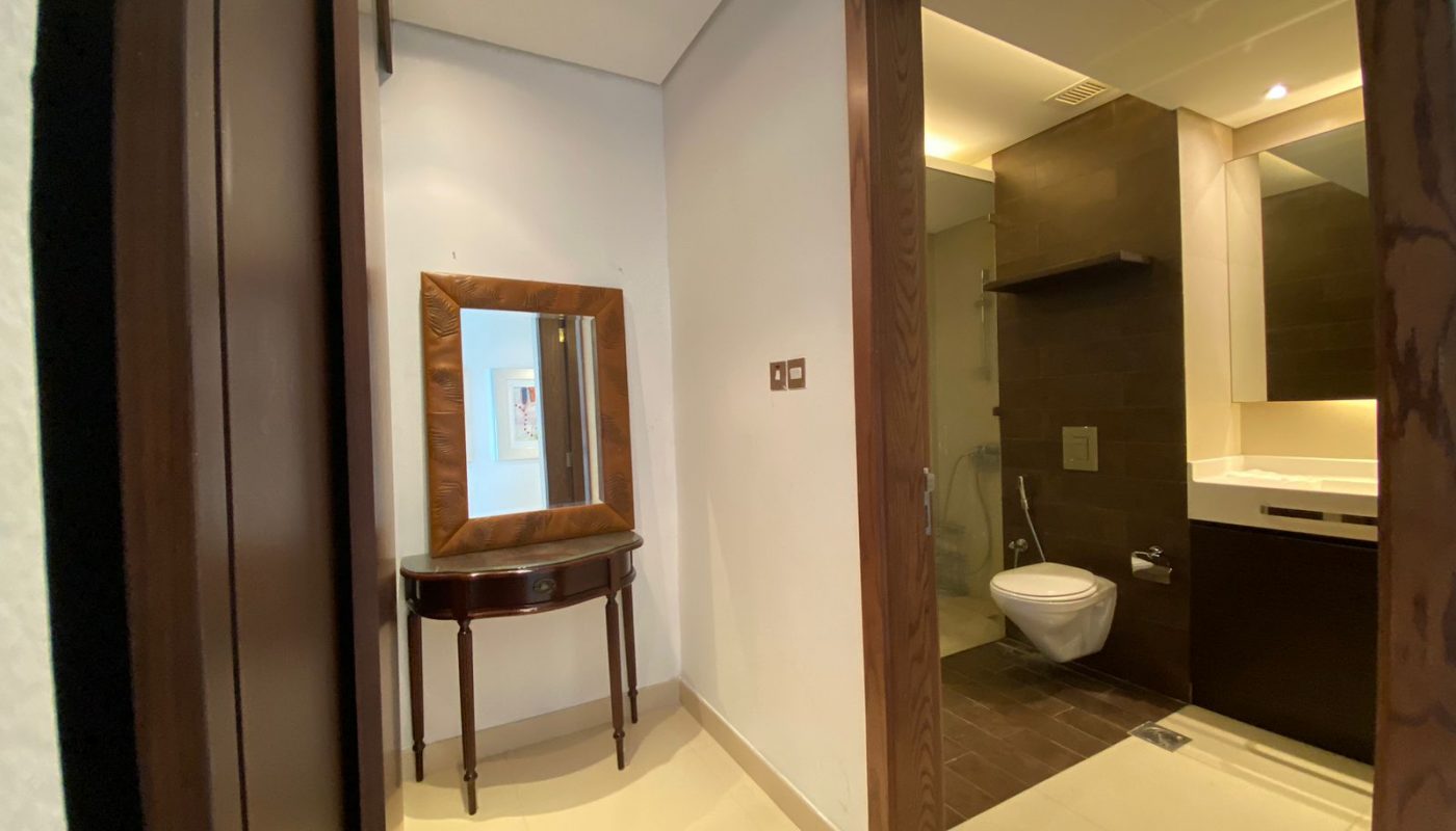 A hallway with a wooden-framed mirror above a table leads to a modern bathroom with a wall-mounted toilet and a sink.