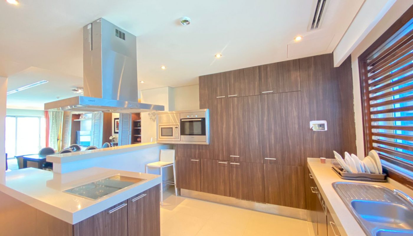 Modern kitchen with wood cabinets, built-in appliances, island with stovetop, double sink, dish rack, and large window with blinds. Bright lighting and an open layout are visible.