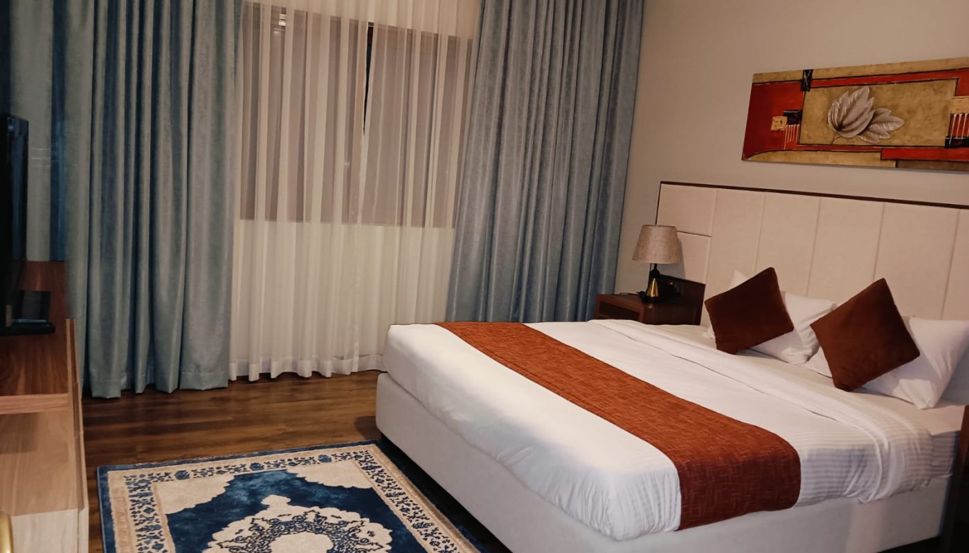 A neatly made double bed with two brown pillows and a matching throw blanket, a bedside lamp, blue curtains, a rug, and wall art in a modern bedroom.