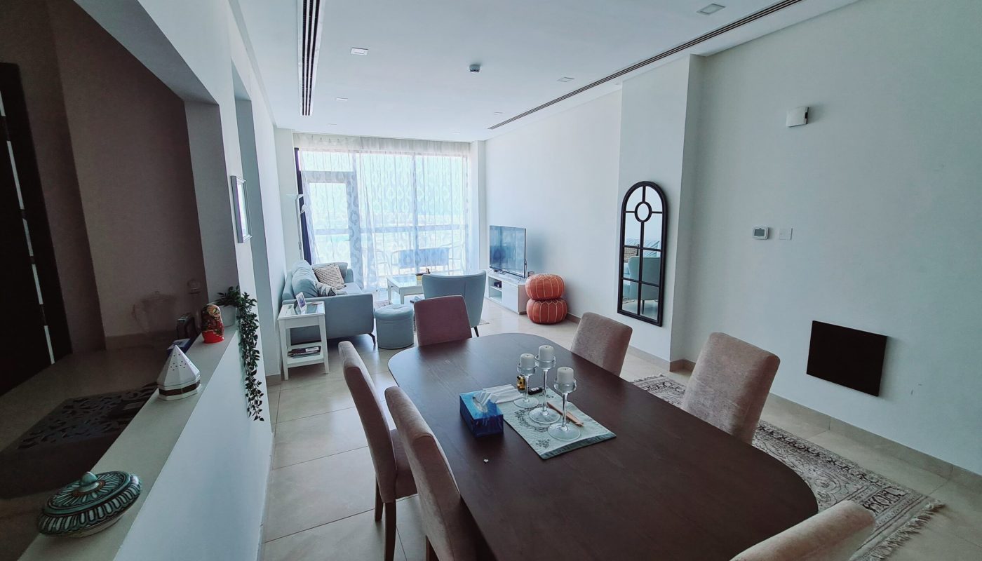 A modern living and dining area with a wooden table, six chairs, a sofa set near a TV, large windows with sheer curtains, and neutral-toned decor.