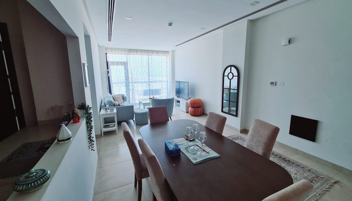 A modern living and dining area with a wooden table, six chairs, a sofa set near a TV, large windows with sheer curtains, and neutral-toned decor.