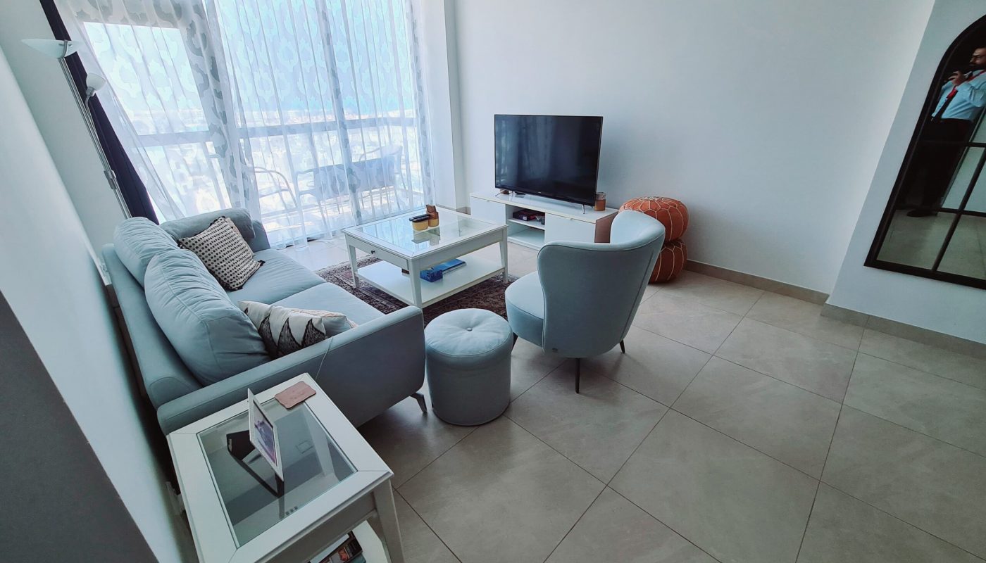 Modern living room with light blue furniture, a TV on a white stand, tiled floor, large window with sheer curtains, and a floor mirror reflecting part of the room.