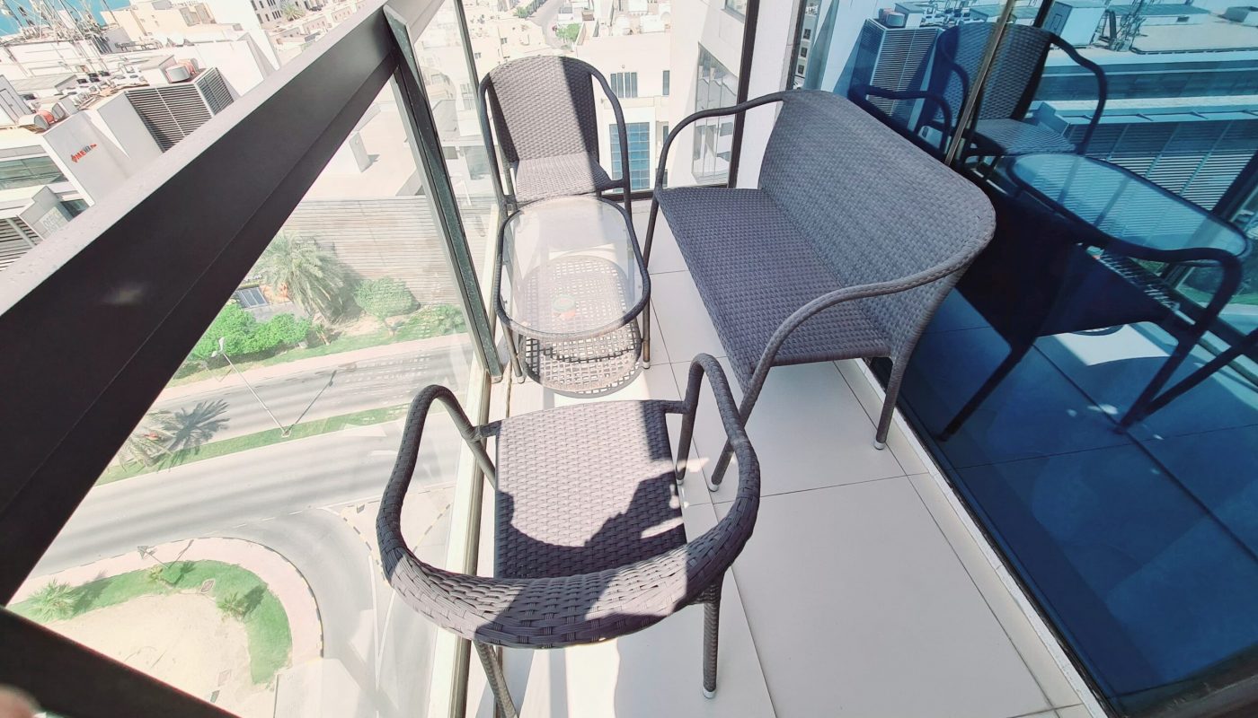 A small balcony with two chairs, a loveseat, and a glass-top table overlooks a street, modern buildings, and the sea in the distance.