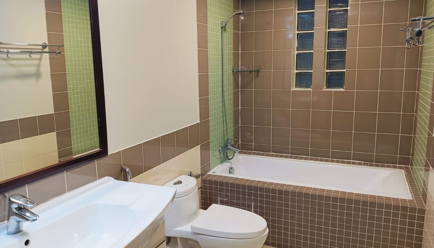 A modern bathroom with a sink, toilet, and bathtub, featuring brown and green wall tiles, a large mirror, and glass block windows above the tub.