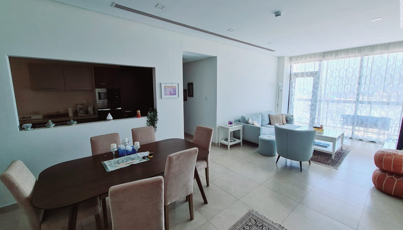 Modern open-plan living and dining area with a wooden table, upholstered chairs, armchairs, large windows with sheer curtains, and a kitchen pass-through window.