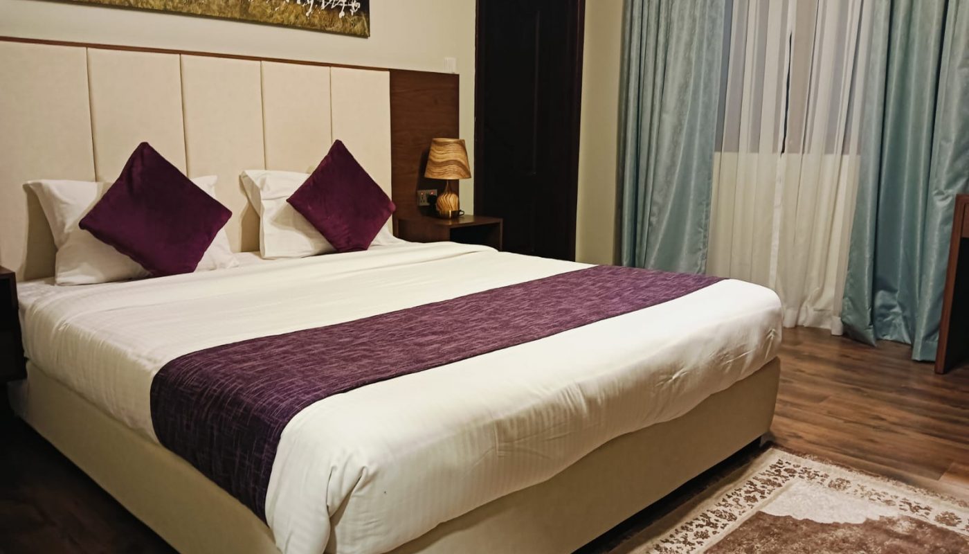 A neatly made bed with white linens and purple accents in a bedroom with wooden flooring, a window with blue curtains, a rug, and a nightstand with a lamp.
