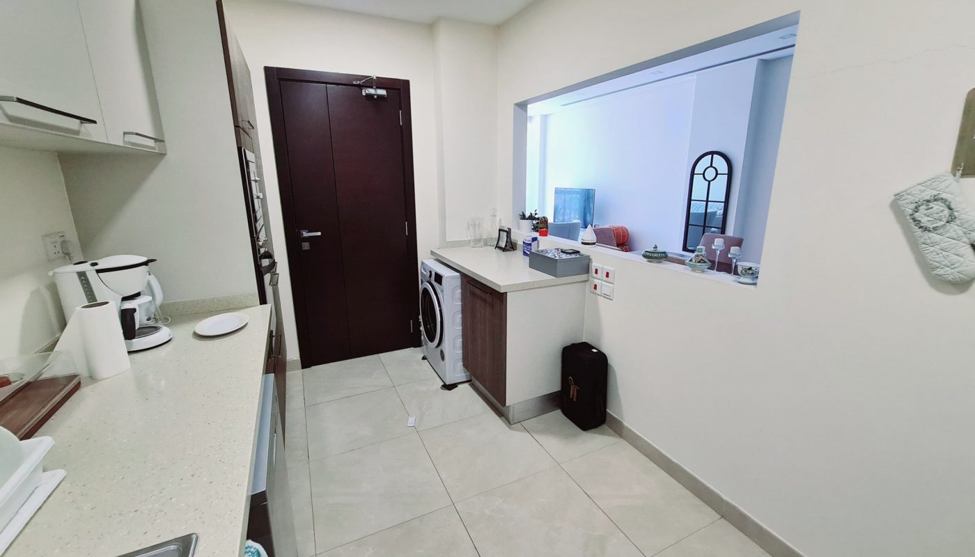 Modern kitchen with white cabinets, appliances on the countertop, a washing machine, and a view into a living area through an open wall window.