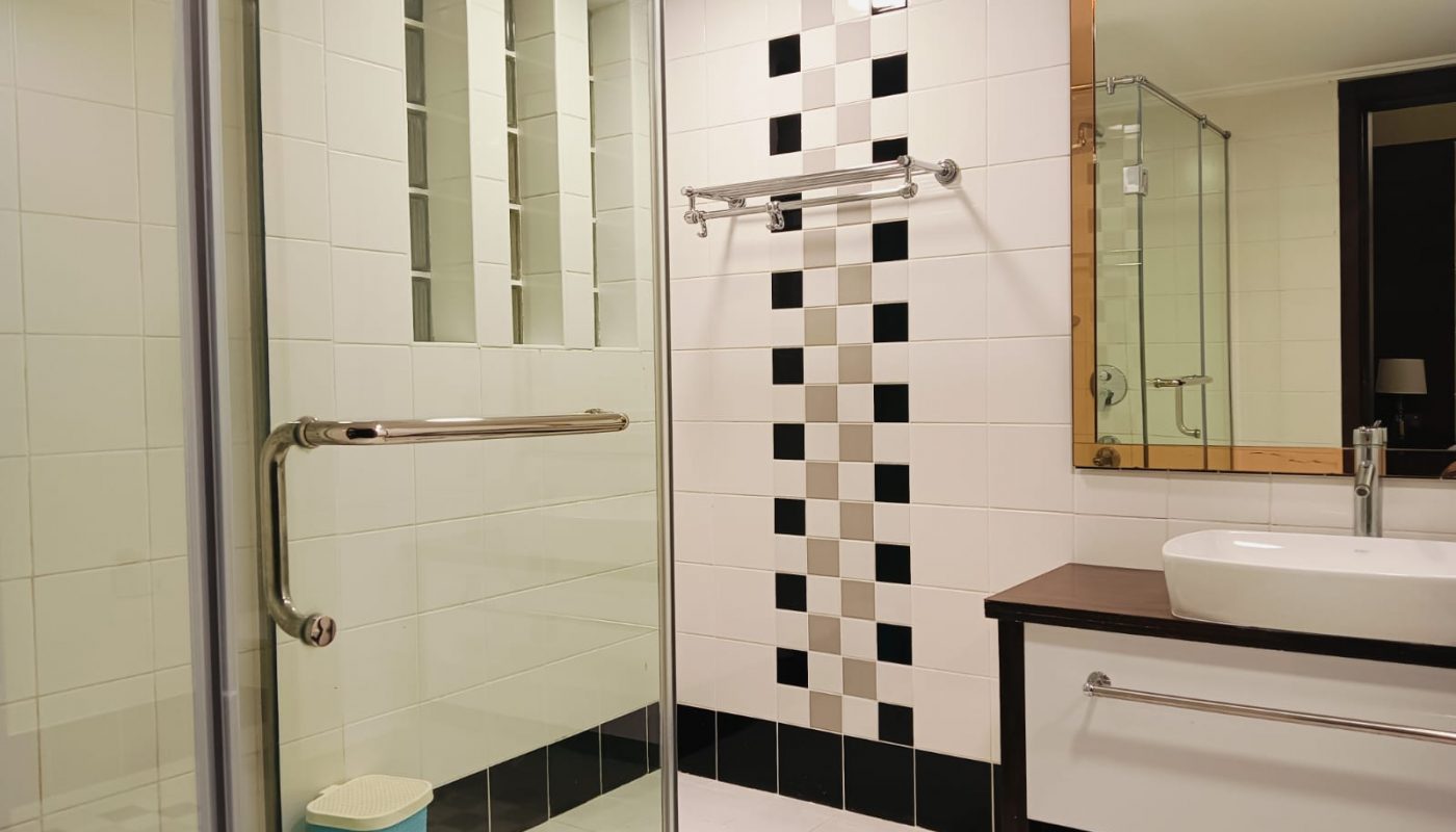 Modern bathroom with glass shower, black and white tiled walls, towel rack, sink with cabinet, large mirror, and a small blue trash bin.