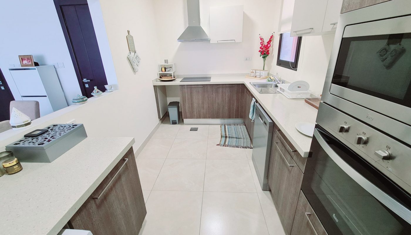 Modern kitchen with light countertops, wooden cabinets, two built-in ovens, a range hood, and a window; a striped rug and various small appliances are visible.