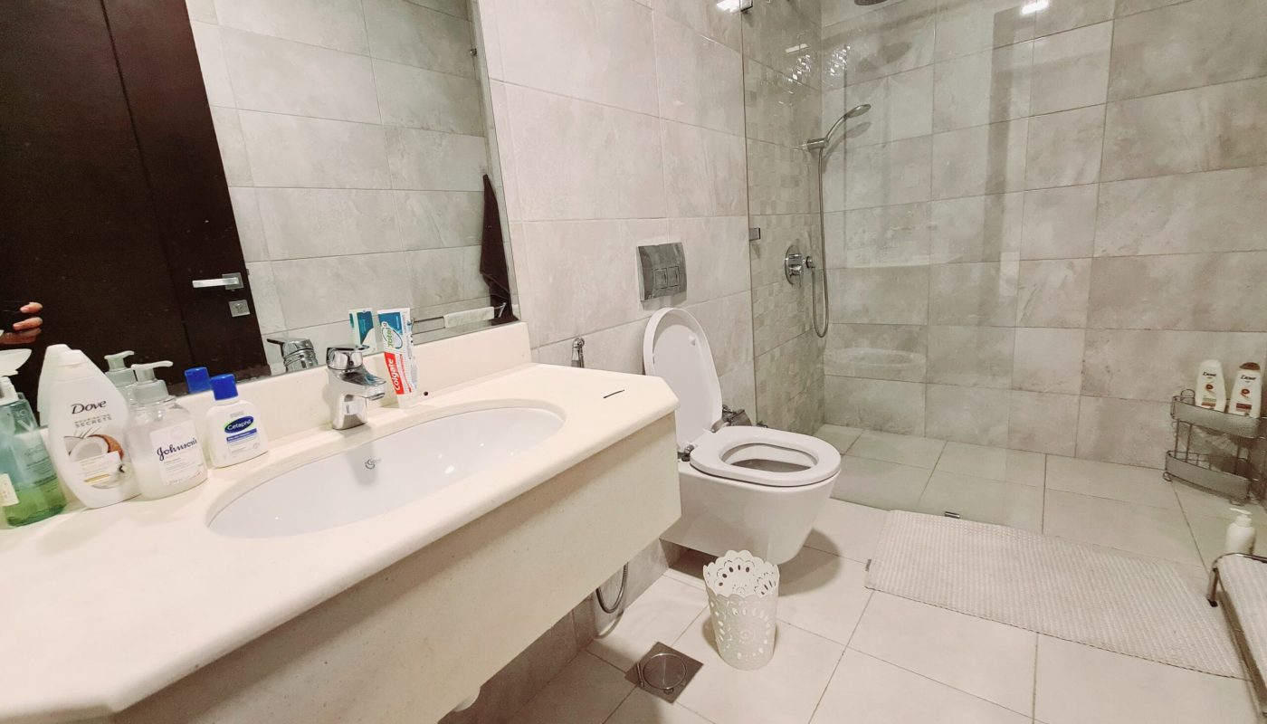 Modern bathroom with a sink, toiletries, wall-mounted toilet, walk-in shower, and toiletries shelf; all surfaces are covered in light gray tiles.
