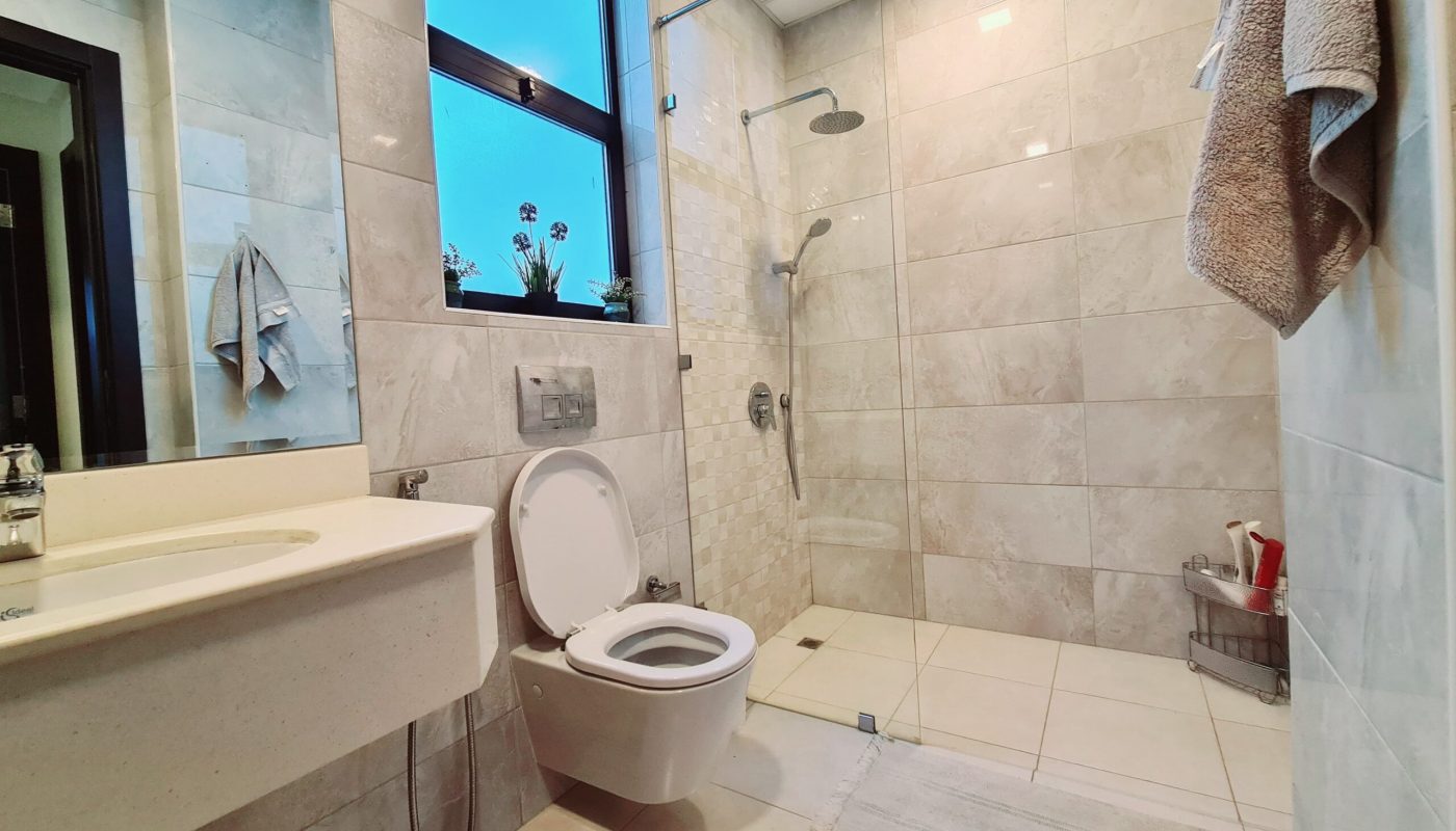 Modern bathroom with beige tiles, wall-mounted toilet, sink, and a glass-enclosed shower area; towels hang on the wall, and a window lets in natural light.