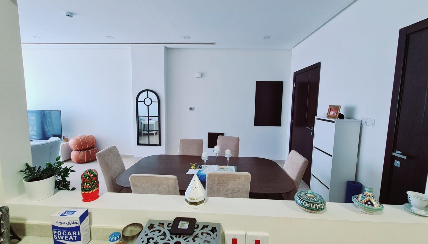 Modern dining room with a wooden table and beige chairs, viewed from a kitchen pass-through. Decorative items and switches are on the counter in the foreground.