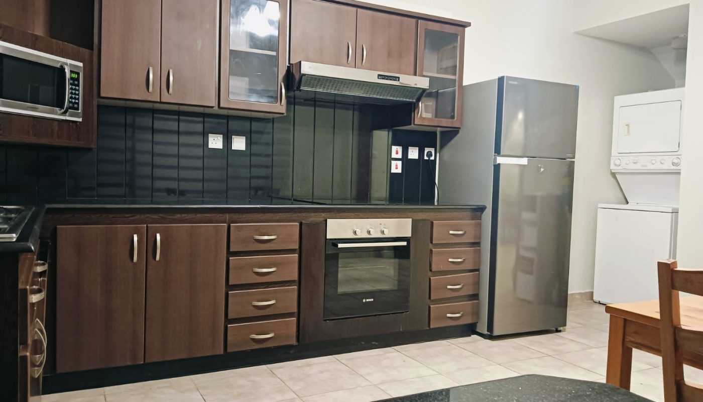 Modern kitchen with dark wood cabinets, built-in oven, electric stove, microwave, stainless steel fridge, and stacked washer-dryer unit; tiled floor and a corner of a wooden table visible.