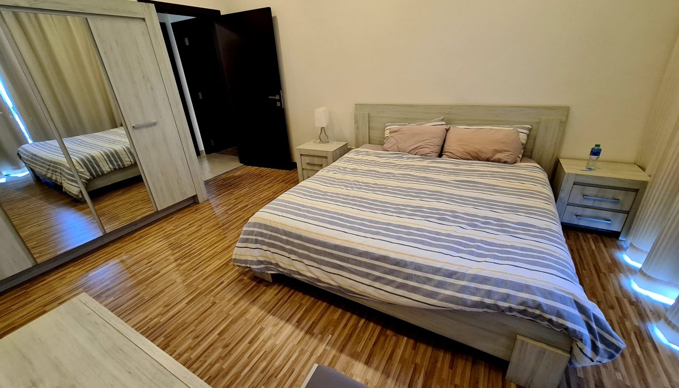 A bedroom with a striped bedspread, two bedside tables, a mirrored wardrobe, wooden flooring, and neutral walls.