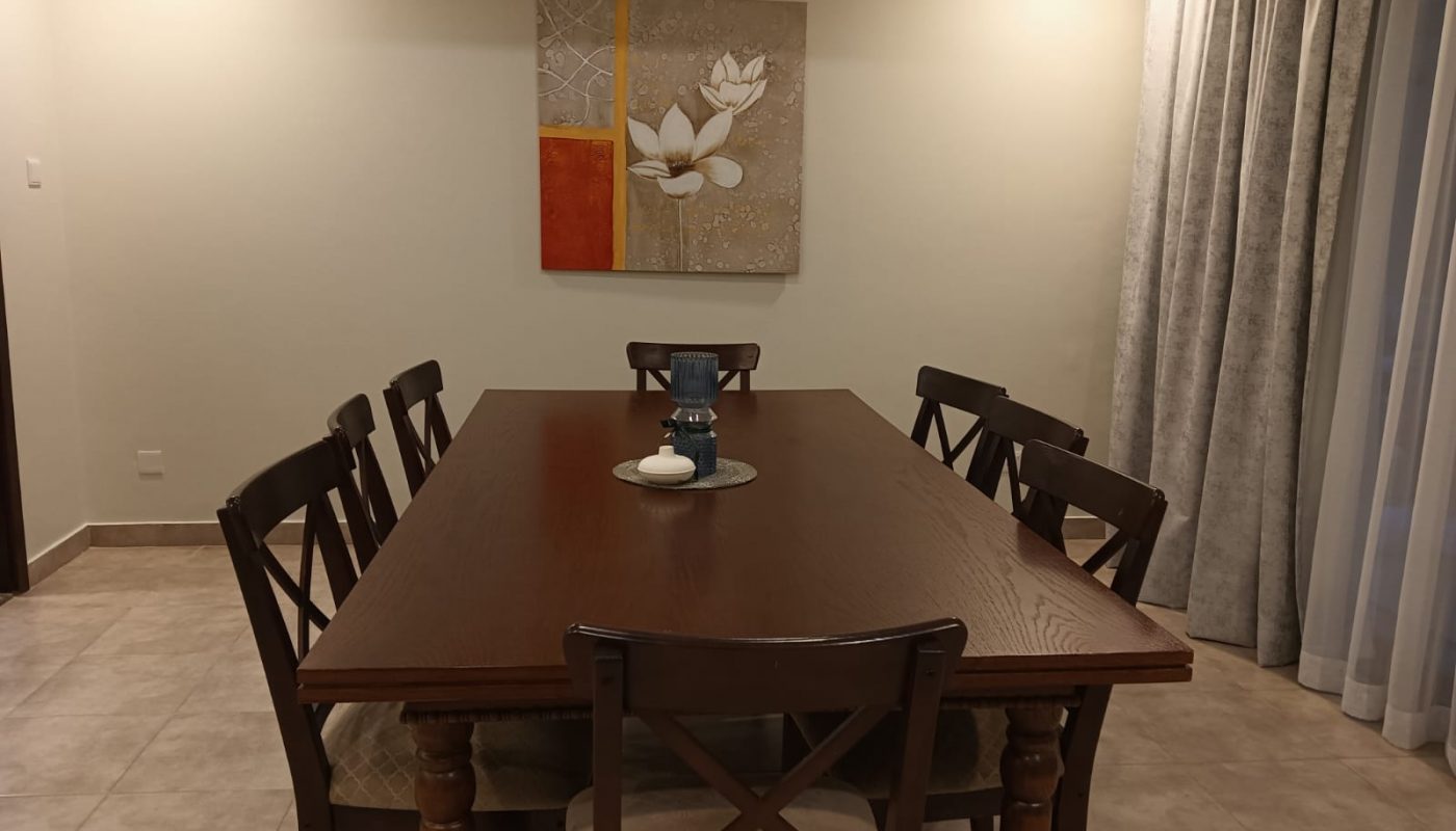 A wooden dining table with six chairs, a decorative centerpiece, tiled floor, light walls, wall art, and curtains on the right side.