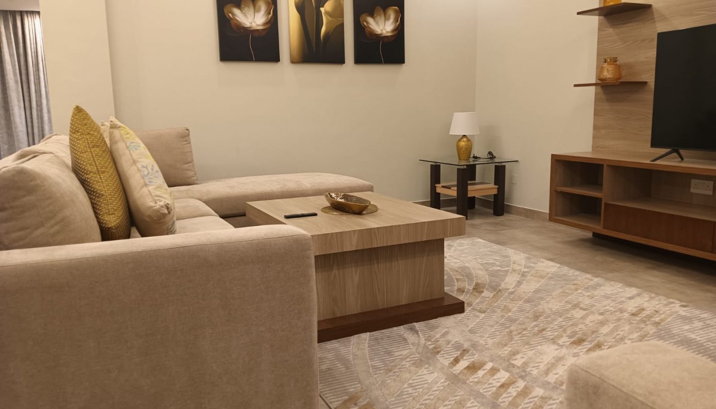 Modern living room with beige sectional sofa, wooden coffee table, patterned rug, wall-mounted TV, decorative shelves, and floral artwork on the wall.