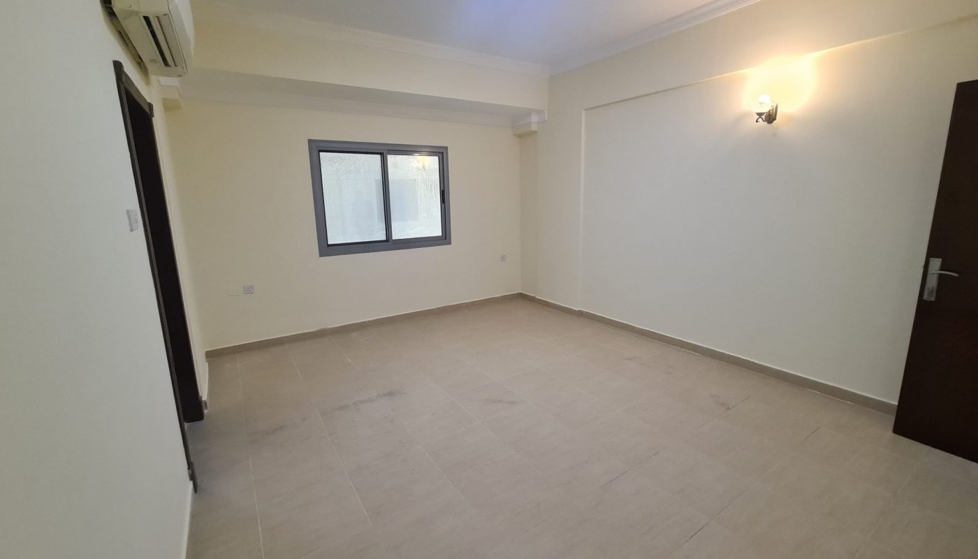 Empty room with beige tiled floor, cream-colored walls, a window, a wooden door, wall-mounted air conditioner, and two light fixtures (one ceiling, one wall).