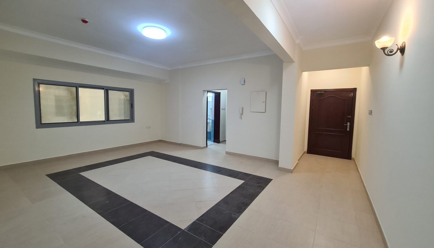 Empty, well-lit room with cream-colored walls, tiled floor featuring a black-bordered square pattern, large window, open doorway, and a closed wooden door.