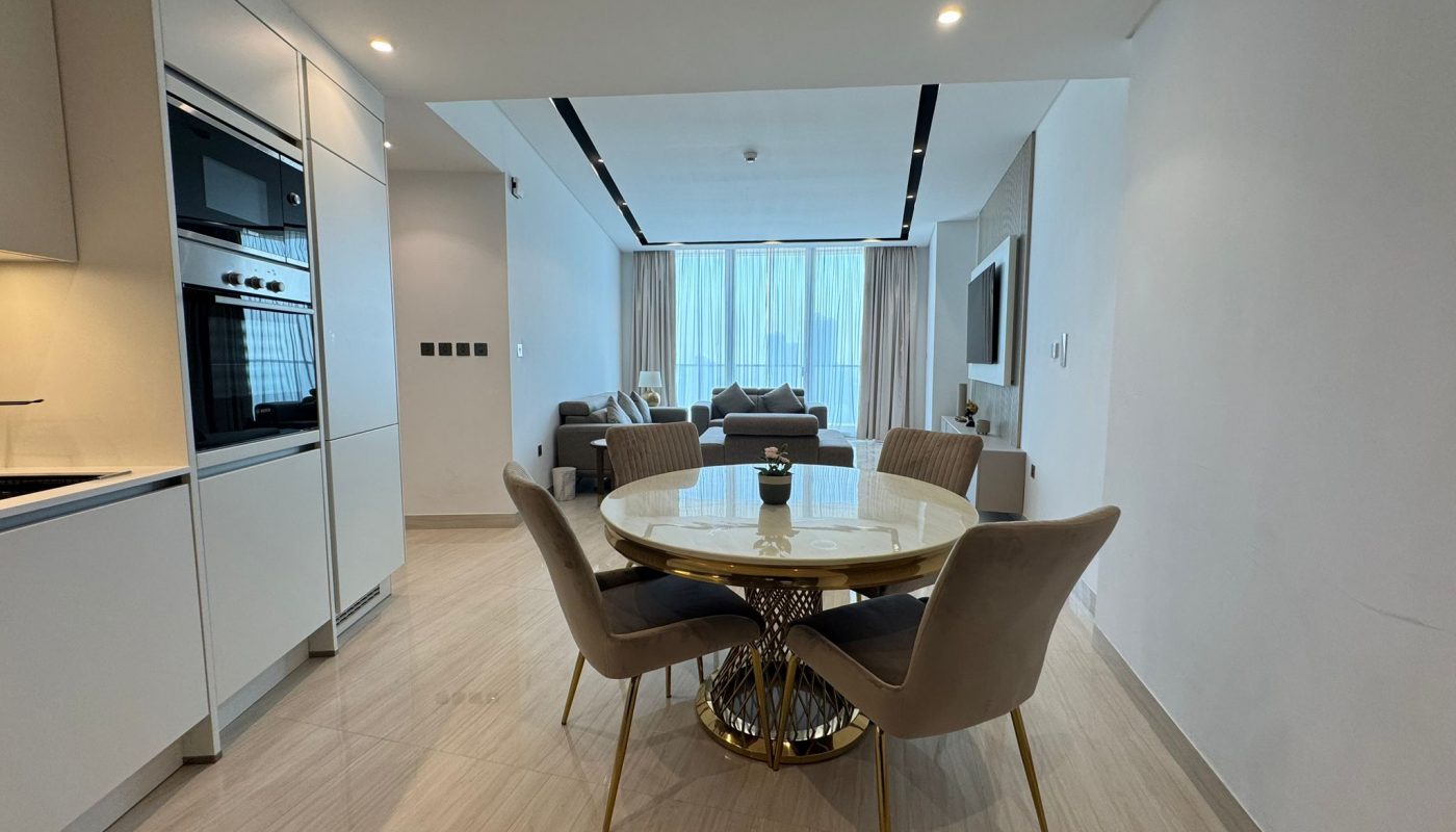 Modern open-plan kitchen and living room with a round dining table and four chairs in the center, light-colored decor, and large windows in the background.