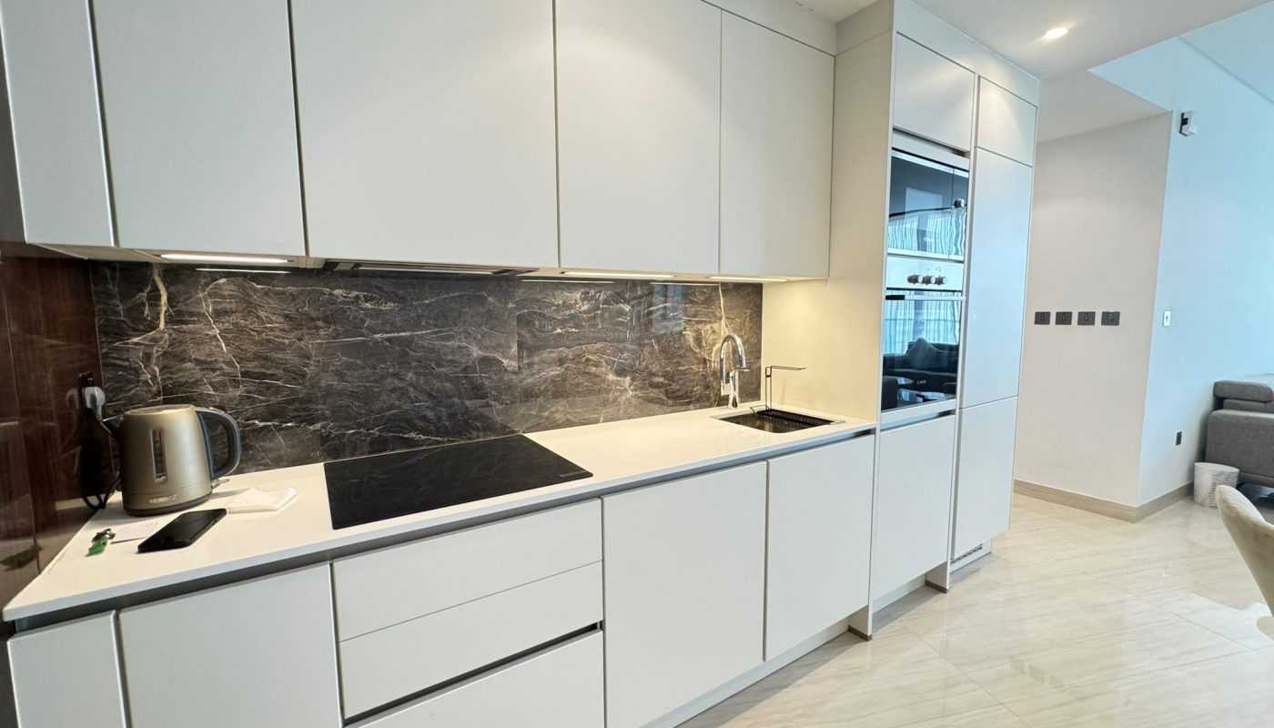Modern kitchen with white cabinets, black marble backsplash, electric cooktop, sink, built-in oven, and a kettle on the counter. Adjacent to a living area with light-colored flooring.