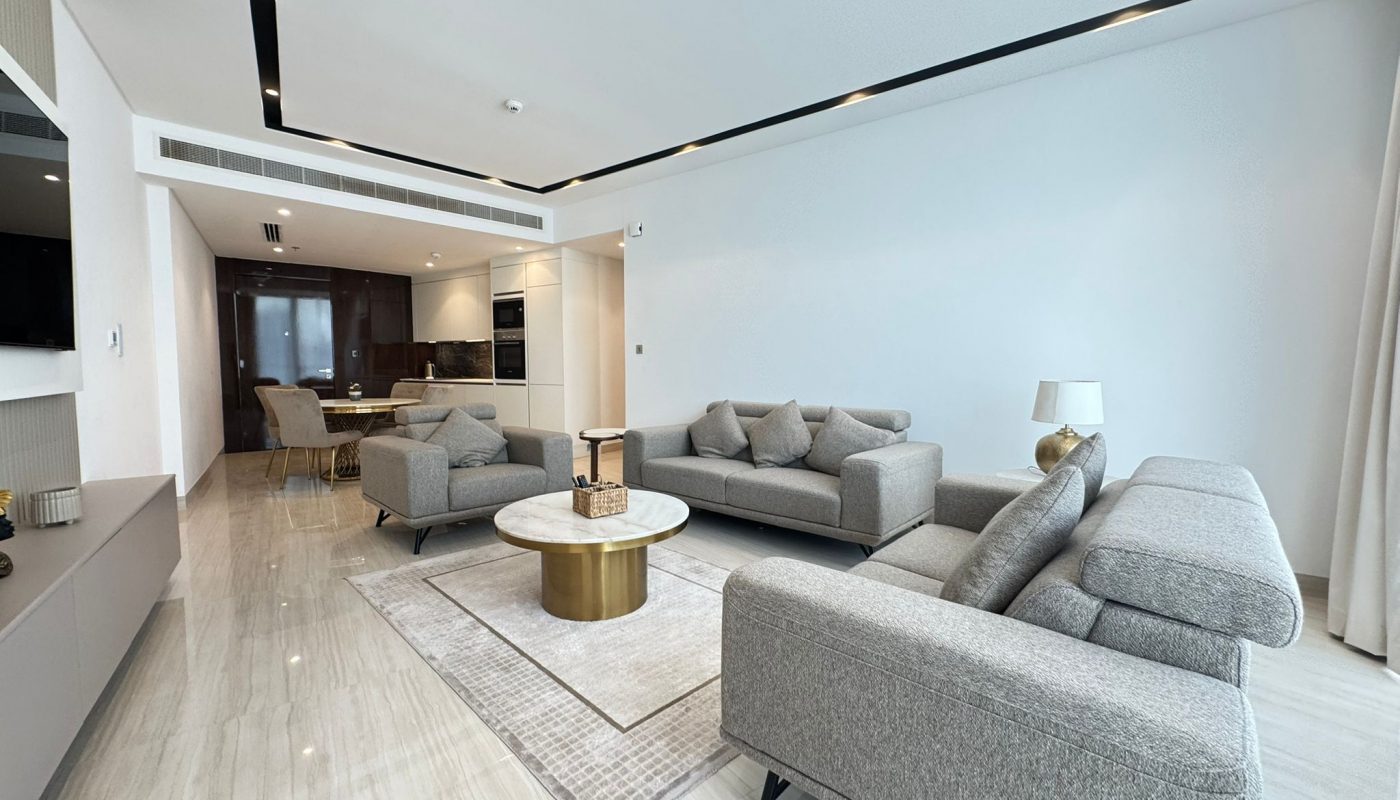 Modern living room with gray sofas, a round gold and white coffee table, a beige rug, and an open kitchen and dining area in the background. The space features light walls and floors.