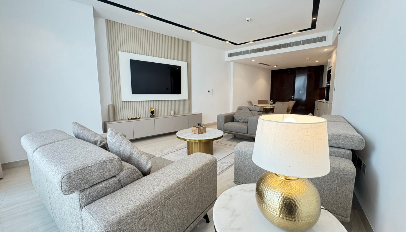 Modern living room with gray sofas, a wall-mounted TV, a round table with a gold lamp, and minimalistic decor in neutral tones.