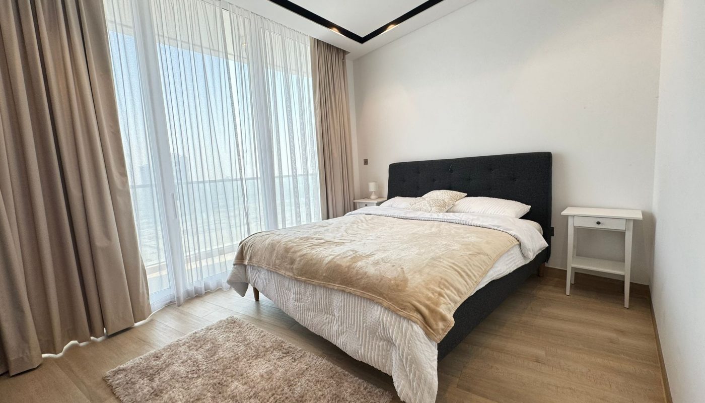 Minimalist bedroom with large window, beige curtains, bed with beige blanket, white nightstand, and a small beige rug on wooden floor.