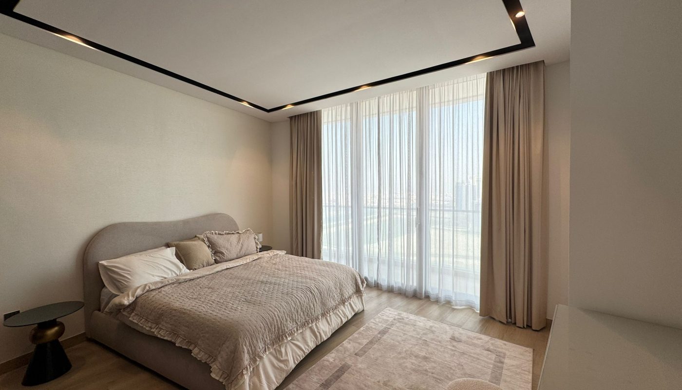 Modern bedroom with a large bed, side table, floor-to-ceiling windows with sheer curtains, a rug, and neutral-toned decor.