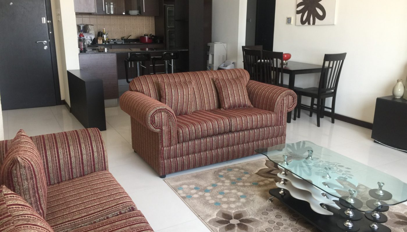 A modern living room with two striped sofas, a glass coffee table, a patterned rug, and an open kitchen and dining area in the background.