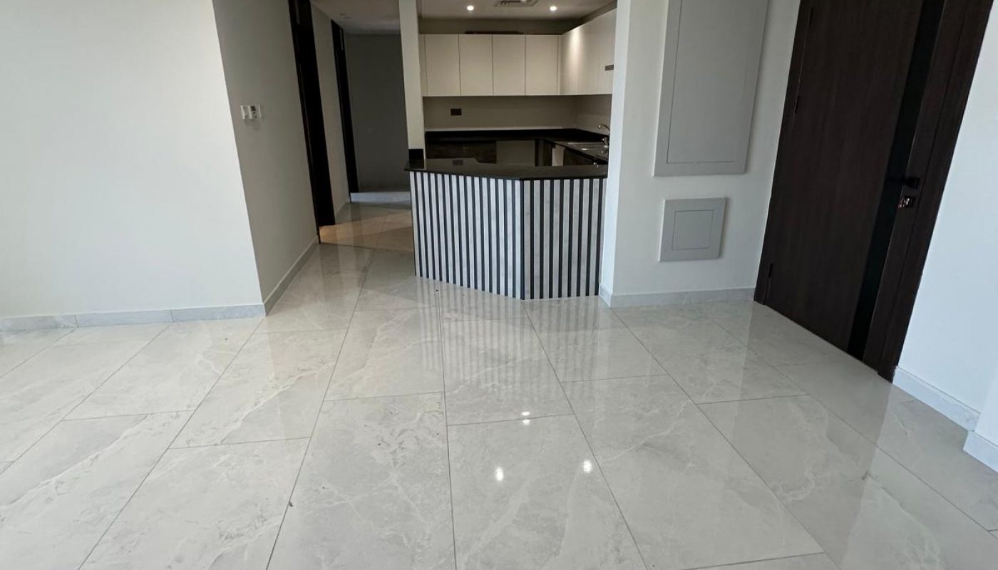 Modern, unfurnished open-plan living area with white marble floors, recessed lighting, and a dark wood entry door leading to a minimalist kitchen with white cabinets and an island.