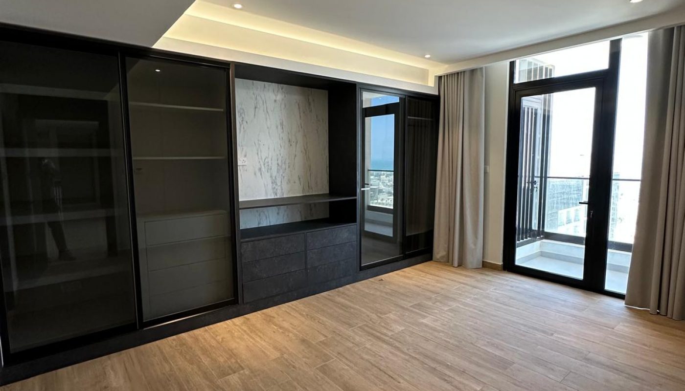 Modern empty room with wood flooring, built-in black shelving units, marble accent wall, recessed lighting, and large windows leading to a balcony.
