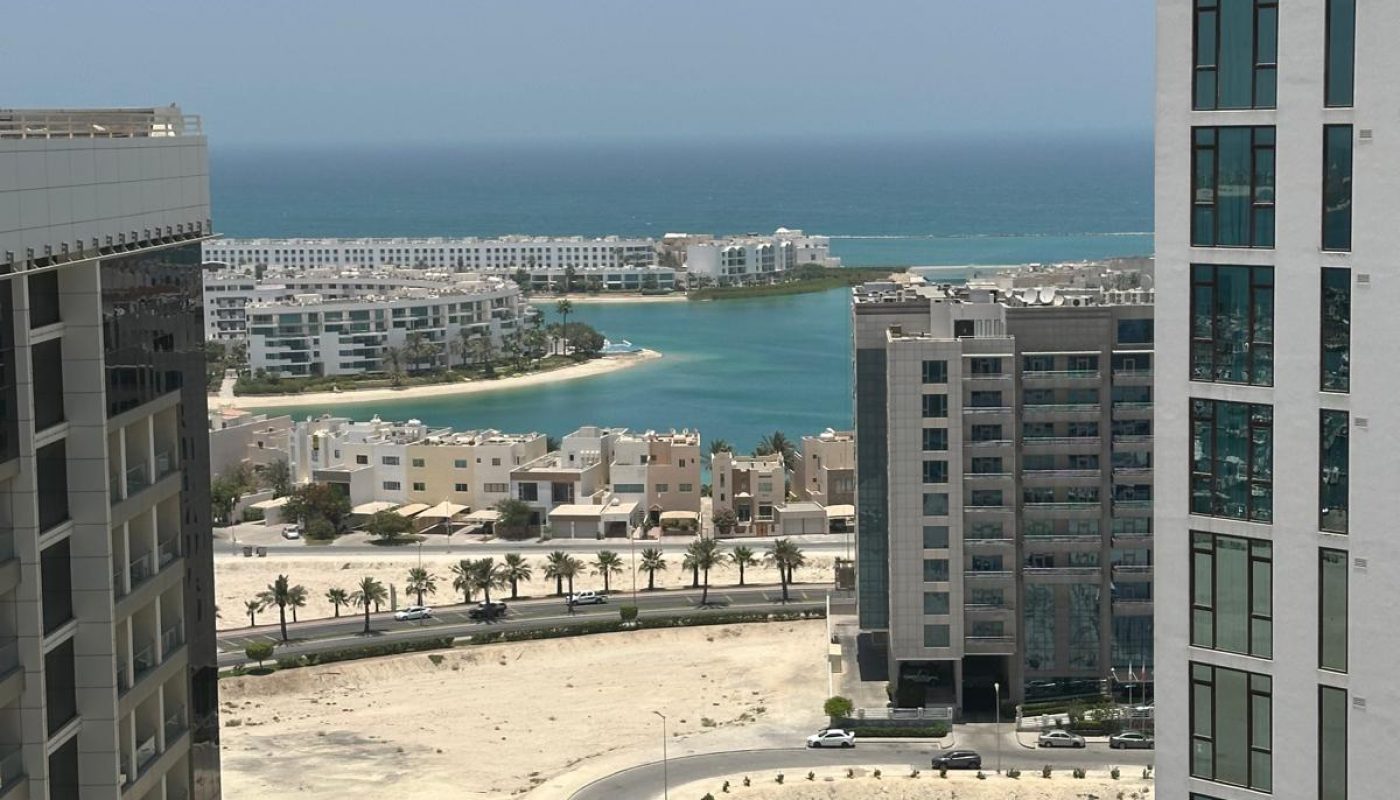 View of tall modern buildings, sandy lots, palm-lined street, and a lagoon leading to the sea under a clear blue sky.