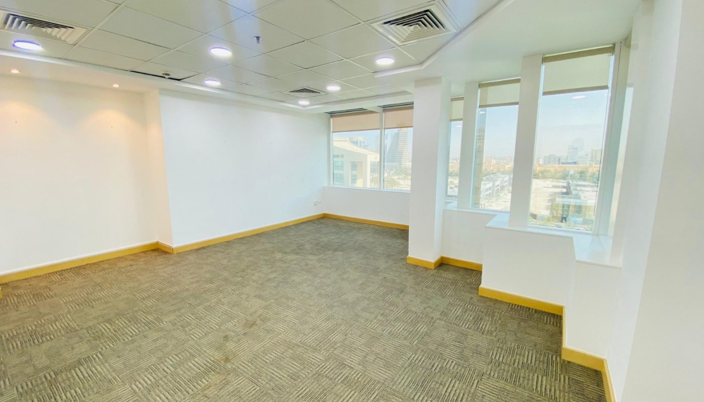 Empty office room with carpeted floor, white walls, ceiling lights, and large windows offering a view of city buildings.