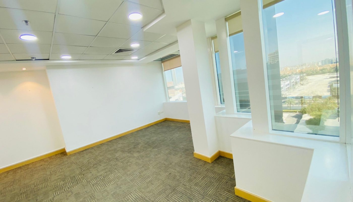Empty office room with white walls, carpeted floor, large windows, and ceiling lights, showing a view of buildings and a parking lot outside.
