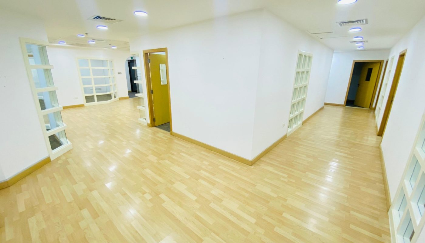 Bright, empty office hallway with light wood flooring, white walls, and several open doorways leading to adjacent rooms. Ceiling lights and glass-panel doors are visible.