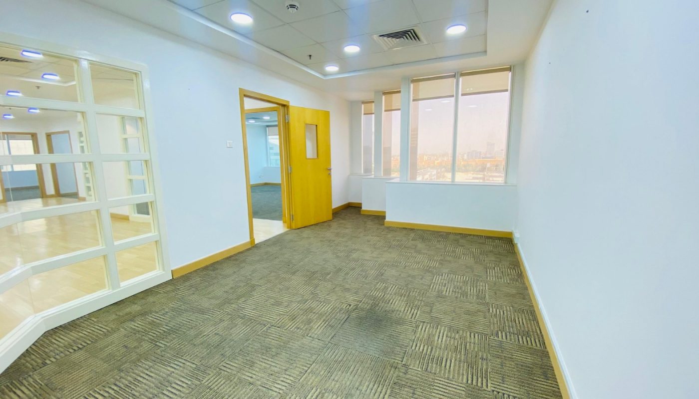 Empty office room with carpeted floor, white walls, and large windows. An open doorway leads to another room with glass partitions and visible overhead lights.