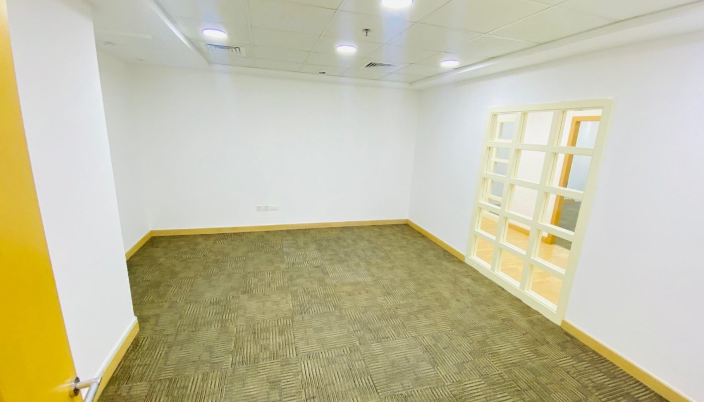 Empty office room with carpeted floor, white walls, ceiling lights, and a glass panel door leading to another room.