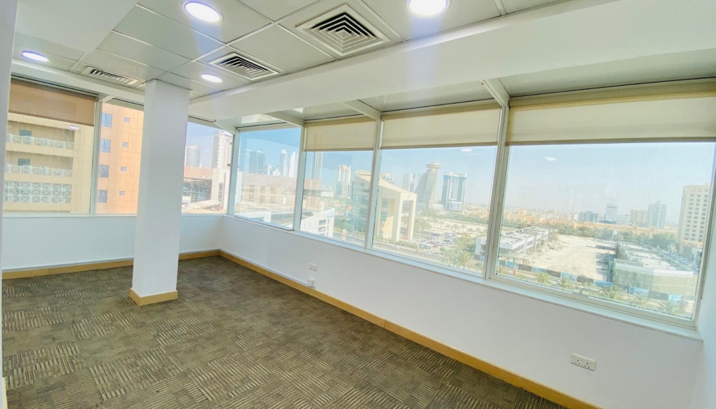 Empty office room with large windows, beige carpet, white walls, and a view of city buildings and streets outside.
