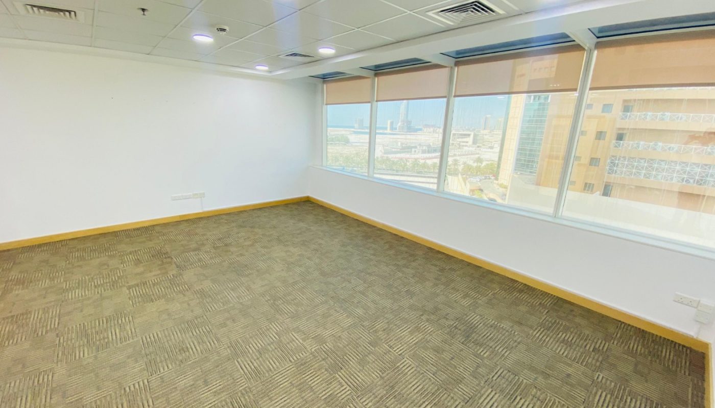 Empty office room with beige carpet, white walls, a drop ceiling with lights, and large windows overlooking a cityscape.