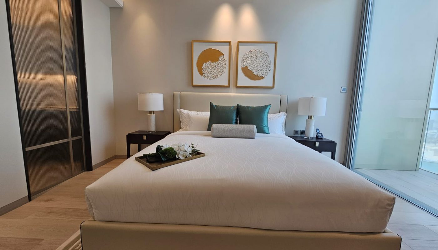 A modern bedroom with a large bed, two nightstands with lamps, decorative pillows, wall art above the headboard, and a floor-to-ceiling window to the right.