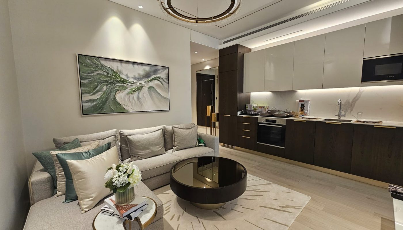 Modern open-plan living room and kitchen with a beige sofa, round glass coffee table, abstract wall art, and a flower vase on a small side table.