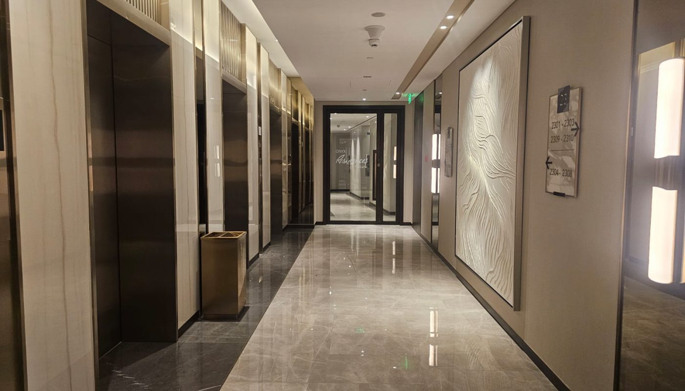 A modern hallway with shiny marble floors, elevators on the left, artwork on the right wall, and a glass door at the end of the corridor.